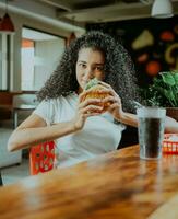 Beautiful latin girl enjoying a delicious hamburger in a restaurant. Portrait of afro girl holding a hamburger in a restaurant. photo