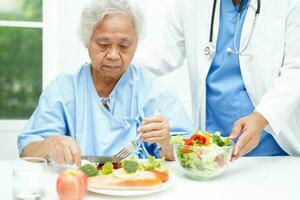 asiático mayor mujer paciente comiendo salmón estaca y vegetal ensalada para sano comida en hospital. foto