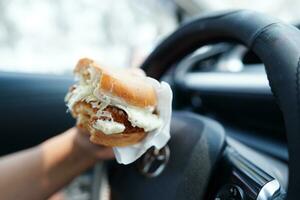 Asian woman driver hold and eat hamburger in car, dangerous and risk an accident. photo