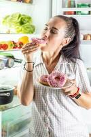 Hungry brunette in pajamas enjoys sweet donuts late at night by the open refrigerator photo