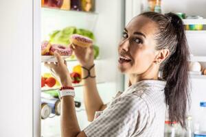 A young hungry woman is about to eat sweet donuts by the open refrigerator late in the evening photo