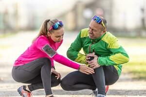 A mature man's knee pain after running is given first aid by a young female runner photo