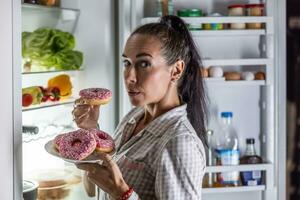 A young hungry woman is about to eat sweet donuts by the open refrigerator late in the evening photo