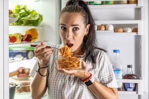 Frightened hungry woman in pajamas is eating spaghetti at the refrigerator at night photo