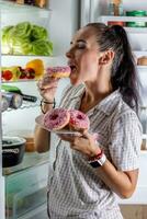 Hungry brunette in pajamas enjoys sweet donuts late at night by the open refrigerator photo