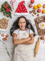 Beautiful dark haired cook blinking laying and on the ground, holding the wooden spoon and being surrounded by gingerbreads, eggs, flour, christmas hat and fruit photo