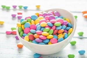 Close up of a white bowl full of smarties with many placed randomly around photo