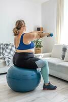 Overweight lady doing sports exercise in living room. He sits on a fitness ball and holds his hands with dumbbells in front of him photo