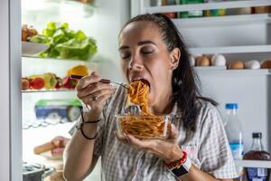 un muy hambriento mujer en pijama es disfrutando espaguetis a el refrigerador a noche foto
