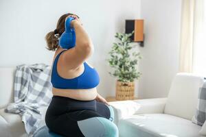 Overweight lady doing sports exercise in living room. She is sitting on a fitness ball and holding a dumbbell on her shoulder photo