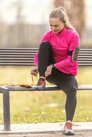 A young woman in sportswear is preparing to run, tying her sneakers on a park bench photo