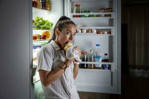 Frightened hungry woman in pajamas is eating cheese at the refrigerator at night photo