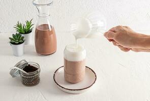 A woman's hand pours whipped milk from a glass jug into coffee in a glass fashionable glass in the form of a beer can. Coffee latte, cappuccino photo