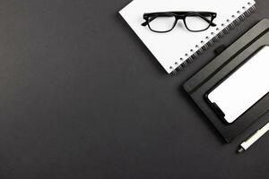 Modern office desk flat lay with smartphone screen mockup, spiral paper notepad and reading glasses on dark background. Top view. Copy space. photo