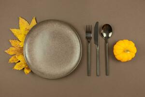 Thanksgiving empty brown plate mockup with fork, knife, spoon, colorful pumpkins and fallen leaves decoration on dark fall background. Autumn flat lay food background. Top view. photo