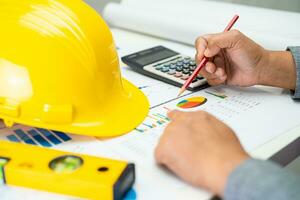 Asian engineer with architectural house plan project blueprint, graph and yellow helmet and engineering construction tools. photo