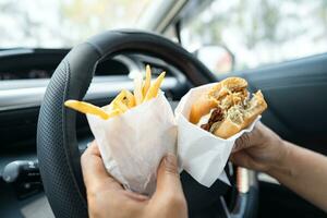 Asian lady holding hamburger and French fries to eat in car, dangerous and risk an accident. photo