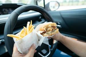 Asian lady holding hamburger and French fries to eat in car, dangerous and risk an accident. photo