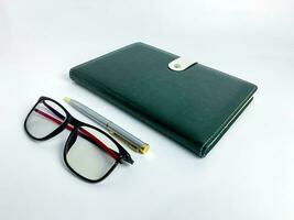Closeup blank green leather diary, pen and glasses on white desk photo