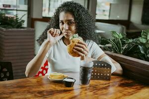 Latin woman sucking her fingers holding a hamburger in a restaurant. Portrait of an afro girl enjoying hamburger in a restaurant photo