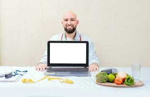 Nutritionist man showing laptop screen. Nutritionist at desk showing blank screen of laptop. Smiling nutritionist showing an advertisement on the laptop photo
