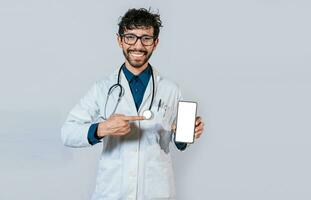 sonriente médico participación y señalando a célula teléfono pantalla. joven médico demostración y señalando a smatphone pantalla aislado foto