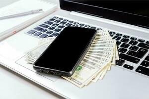 Cell phone on laptop keyboard and dollar bills. Mobile phone on top of dollar bills on laptop keyboard. Top view of laptop with cell phone and dollar bills photo