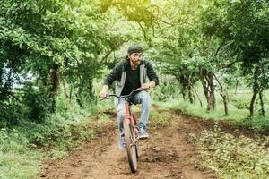 Adolescente chico montando un bicicleta en el campo, retrato de un chico en gorra montando un bicicleta en un país camino, persona montando un bicicleta en el campo, ciclista persona en su bicicleta en un país la carretera bosque foto