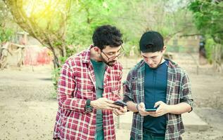 Two teenage guys pointing and checking their cell phones, Two friends looking at the content of their cell phone. Friend showing cell phone to his friend, Smiling friends checking cell phones photo