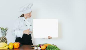 Chef woman with vegetables at table holding blank menu. Girl chef in the kitchen showing a blank board, Beautiful woman chef with table of vegetables holding a blank board photo