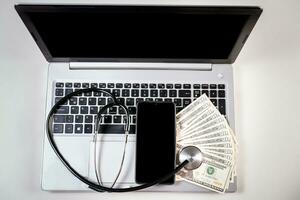 Mobile phone with dollar bills and stethoscope on laptop keyboard. Stethoscope on top of dollar bills on laptop keyboard photo