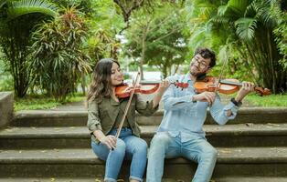 Portrait of two young violists playing violin sitting on stairs outside. Portrait of smiling violinists sitting on stairs playing violin. Man and woman sitting on stairs playing violin photo