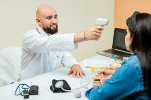 Doctor measuring temperature with infrared gun to female patient, Doctor taking temperature to woman patient in office, Male doctor taking temperature with gun to patient photo