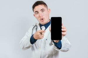 Amazed young doctor pointing cell phone screen isolated. Latin doctor showing an app on cell phone screen. photo
