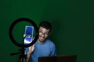 estudiante recepción en línea clases hombre recepción en línea clases con teléfono. chico con ordenador portátil haciendo vídeo Blog en teléfono. en línea clases concepto. chico en un vídeo llamada trabajando desde hogar foto