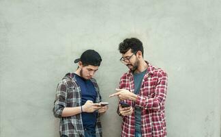 Two young friends leaning against a wall checking their cell phones, Two friends leaning against a wall looking at the content on their phones. Friend showing cell phone to his friend outdoors photo