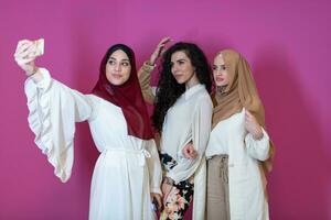 Group of beautiful muslim women two of them in fashionable dress with hijab using mobile phone while taking selfie picture isolated on pink background representing modern islam fashion technology photo