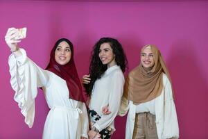 Group of beautiful muslim women two of them in fashionable dress with hijab using mobile phone while taking selfie picture isolated on pink background representing modern islam fashion technology photo