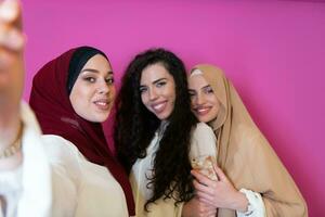 Group of beautiful muslim women two of them in fashionable dress with hijab using mobile phone while taking selfie picture isolated on pink background representing modern islam fashion technology photo