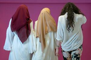 group portrait of beautiful muslim women two of them in fashionable dress with hijab isolated on pink background representing modern islam fashion and ramadan kareem concept photo