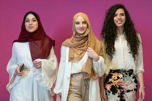 Group portrait of beautiful muslim women two of them in fashionable dress with hijab isolated on pink background representing modern islam fashion and ramadan kareem concept photo