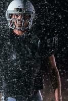 americano fútbol americano campo solitario atleta guerrero en pie en un campo sostiene su casco y Listo a jugar. jugador preparando a correr, ataque y Puntuación aterrizaje. lluvioso noche con dramático niebla, azul ligero foto