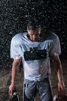 American Football Field Lonely Athlete Warrior Standing on a Field Holds his Helmet and Ready to Play. Player Preparing to Run, Attack and Score Touchdown. Rainy Night with Dramatic Fog, Blue Light photo