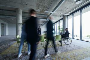 A businessman in a wheelchair sits at a table in a large, modern office, while his colleagues gather around, their steps blurred, symbolizing inclusivity, support, and unity in the face of challenges. photo