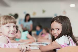 Little girls sitting in elementary school drawing on paper with their friends while sitting in a modern classroom photo