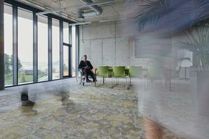 A businessman in a wheelchair sits at a table in a large, modern office, while his colleagues gather around, their steps blurred, symbolizing inclusivity, support, and unity in the face of challenges. photo