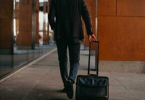 ir a la terminal del aeropuerto. viajero de negocios confiado caminando por las calles de la ciudad y tirando de su maleta bebiendo café y hablando en el teléfono inteligente foto