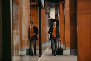 Going to airport terminal. Confident businessman traveler walking on city streets and pulling his suitcase drinking coffee and speaking on smartphone photo