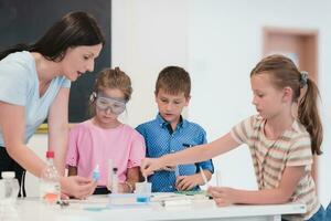 Elementary School Science Classroom Enthusiastic Teacher Explains Chemistry to Diverse Group of Children, Little Boy Mixes Chemicals in Beakers. Children Learn with Interest photo