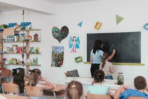 elemental escuela. el hembra profesor Ayudar el niño estudiante mientras escritura el responder en el pizarra foto